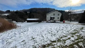 Weißes Wohnhaus mit schneebedecktem Garten und Hügellandschaft im Hintergrund unter blauem Himmel.
