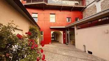 Außenansicht eines modernen Gebäudes mit roter Fassade, Balkon und gepflastertem Innenhof mit Rosen.