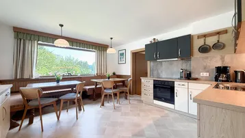Gemütliche Wohnküche mit Essbereich, Fliesenboden und Blick ins Grüne durch ein großes Fenster.