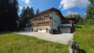 Großes Gebäude mit mehreren Balkonen, umgeben von grüner Natur und Bergen unter blauem Himmel.