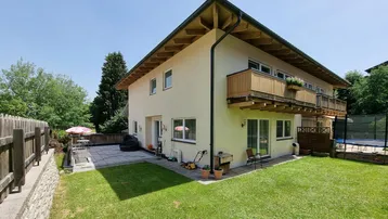 Hausfassade mit gepflegtem Rasen, großer Terrasse und Balkon unter blauem Himmel.