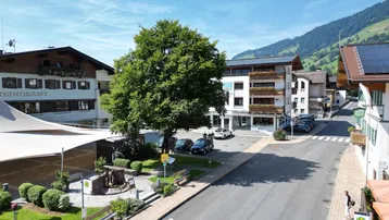 Luftaufnahme einer belebten Dorfstraße mit traditionellen Gebäuden, Bäumen und Bergen im Hintergrund unter blauem Himmel.
