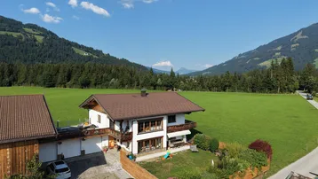 Luftaufnahme eines charmanten Hauses mit Holzbalkonen, umgeben von grünen Wiesen und einer malerischen Berglandschaft.