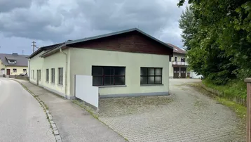 Außenansicht eines Gewerbegebäudes mit gepflastertem Vorplatz unter bewölktem Himmel und umgebender Natur.