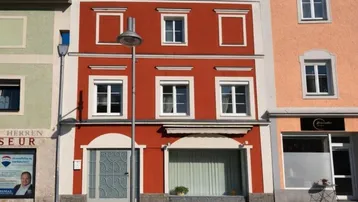 Rotes Gebäude mit weißer Fassadenverzierung und Geschäften im Erdgeschoss an einem sonnigen Tag.