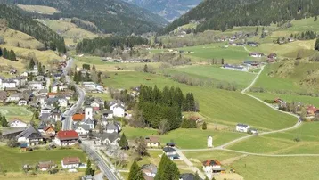 Weitläufige Luftaufnahme eines Dorfes in einem grünen Tal, umgeben von bewaldeten Bergen.