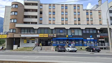 Gebäudeansicht mit Geschäftsflächen im Erdgeschoss und Wohnungen darüber, umgeben von einer städtischen Straße.