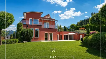 Moderne Villa mit roter Fassade, großem gepflegten Rasen und umgebender grüner Landschaft unter blauem Himmel.