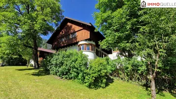 Hausfassade mit Holzelementen und einem gepflegten Garten umgeben von Bäumen.