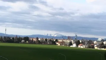 Weitläufiger Blick über eine Stadt mit grünen Feldern und schneebedeckten Bergen im Hintergrund.