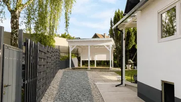 Seitlicher Zugang zum Haus mit Kiesweg, gepflegtem Garten und einer Pergola im Hintergrund.