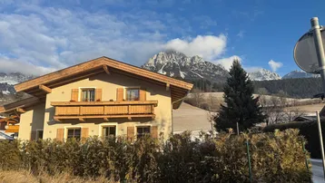 Alpines Haus mit Holzbalkon und beeindruckendem Bergblick unter blauem Himmel.