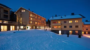Mehrere moderne Gebäude in einer verschneiten Berglandschaft bei Dämmerung mit beleuchteten Fenstern.