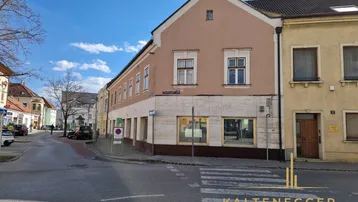 Gebäude an einer Straßenecke mit hellbrauner Fassade und großen Fenstern im Erdgeschoss.