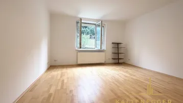 Heller Wohnraum mit Parkettboden, weißen Wänden und Ausblick ins Grüne durch das Fenster.