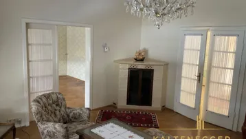 Helles Wohnzimmer mit Kronleuchter, Kachelofen und Parkettboden, offen zu einem weiteren Raum.