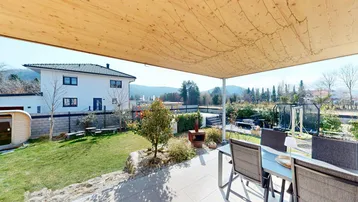 Überdachte Terrasse mit Esstisch und Stühlen, umgeben von einem weitläufigen Garten und Bergblick.