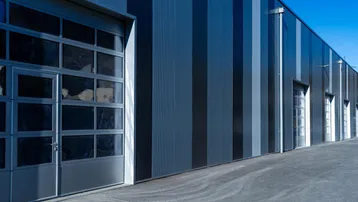 Moderne Industriefassade mit vertikalen Paneelen und großen Glas-Sektionaltoren, die viel Licht hereinlassen.