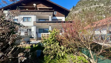 Mehrstöckiges Wohnhaus mit Gartenbereich und Balkonen, umgeben von grüner Natur und Bergen.