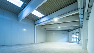 Geräumige, leere Lagerhalle mit hohen Decken, Betonboden und Oberlichtern für natürliches Licht.