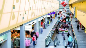 Helles Einkaufszentrum mit mehreren Ebenen, Rolltreppen und Geschäften, beleuchtet durch ein großes Glasdach.