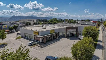 Luftaufnahme eines Gewerbegebäudes mit großem Parkplatz und angrenzender Straße, umgeben von Bäumen und Bergen im Hintergrund unter blauem Himmel.