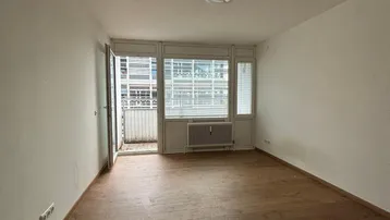 Helles Zimmer mit Holzboden, weißen Wänden und Zugang zu einem Balkon.