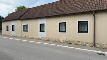 Längliche Hausfassade mit beiger Putzfarbe, braunem Ziegeldach und dunklen Fenstern an einer ruhigen Straße.