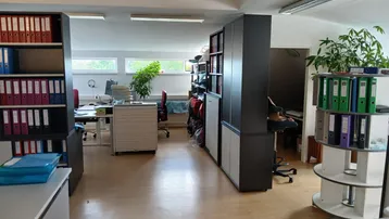 Großes, offenes Büro mit mehreren Arbeitsplätzen, Regalen und Fenstern, die viel Tageslicht hereinlassen.