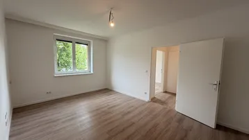 Helles, leeres Zimmer mit Laminatboden und Blick ins Grüne durch das Fenster.