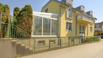 Gelbe Hausfassade mit Wintergarten und Außentreppe, umgeben von grünen Büschen und einem Zaun.
