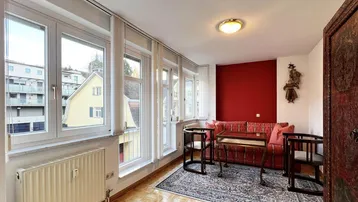 Helles Wohnzimmer mit roter Akzentwand, Parkettboden und vielen Fenstern mit Ausblick.
