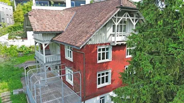 Historisches Gebäude mit roter Fassade, Ziegeldach und einem Balkon, umgeben von Bäumen.