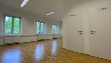 Geräumiger, heller Büroraum mit Parkettboden, weißen Wänden und mehreren Fenstern mit Ausblick.