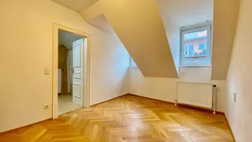 Gemütliches Zimmer mit Fischgrätparkett, Dachschrägen und Fenster mit Blick nach draußen.