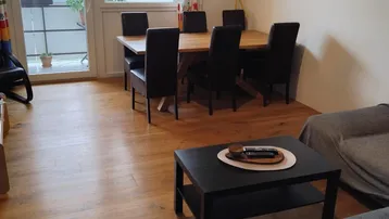 Helles Wohn- und Esszimmer mit Holzboden, Zugang zum Balkon und Blick ins Grüne.