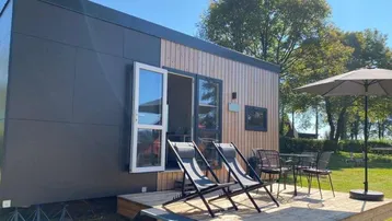 Modernes kleines Haus mit Holzterrasse, Sitzgelegenheiten und Sonnenschirm im Grünen.