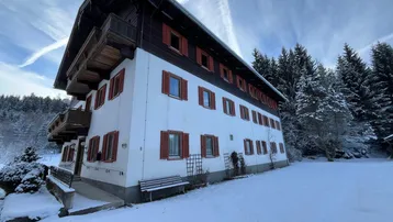 Traditionelles alpines Gebäude mit roten Fensterläden und Holzbalkonen, umgeben von verschneiten Bäumen und Landschaft.