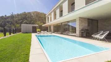 Modernes Haus mit großem Swimmingpool, weitläufigem Rasen und Bergblick unter strahlendem Himmel.
