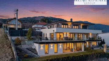 Moderne Villa mit mehreren Terrassen und Balkonen, eingebettet in eine Hanglage mit Bergblick bei Sonnenuntergang.