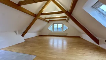 Helles Dachgeschosszimmer mit sichtbaren Holzbalken, Dachschrägen und einem besonderen Giebelfenster.
