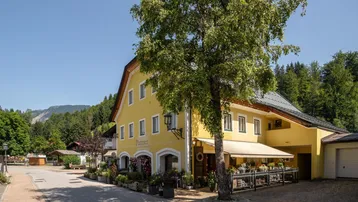 Traditionelles gelbes Gebäude mit grünen Fensterläden und einer Markise, umgeben von üppiger Natur und Bergen.