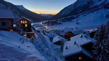 Winterliche Abendstimmung in einem verschneiten Bergdorf mit beleuchteten Häusern und Straßen.