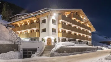 Modernes Gebäude im alpinen Stil mit beleuchteten Balkonen und verschneiter Umgebung bei Nacht.