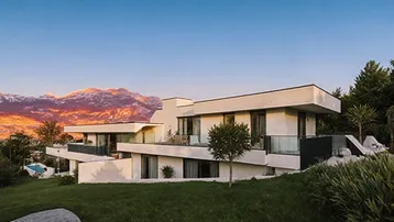 Moderne Villa mit mehreren Ebenen und Terrassen, umgeben von grüner Landschaft bei Sonnenuntergang.