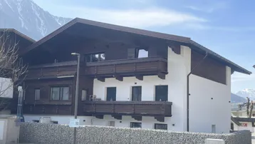 Traditionelles Gebäude mit Holzbalkonen und weißer Fassade vor alpiner Bergkulisse.