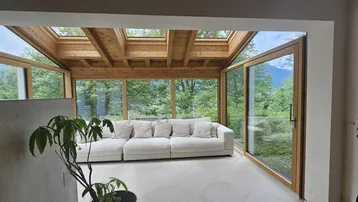 Heller Wintergarten mit bequemer Couch, Holzbalken und weitem Blick ins Grüne.
