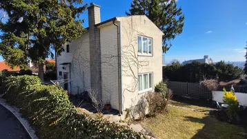 Seitenansicht des Hauses mit Gartenbereich, umgeben von Bäumen und Hecken unter blauem Himmel.