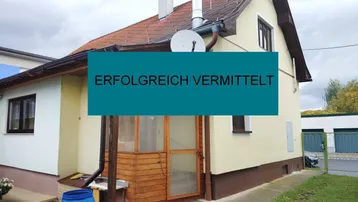 Kleines Einfamilienhaus mit Ziegeldach, heller Fassade und hölzernem Eingangsbereich, umgeben von Grünflächen.