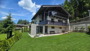 Modernes Haus mit großem Garten, Glasanbau und Balkon in alpiner Umgebung.